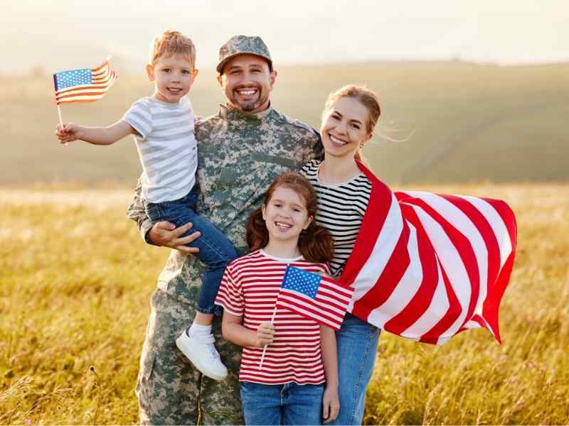 U.S. MILITARY MAN SMILING AND POSING FOR PHOTO WITH WIFE, DAUGHTER, SON, AND AMERICAN FLAGS IN A FIELD