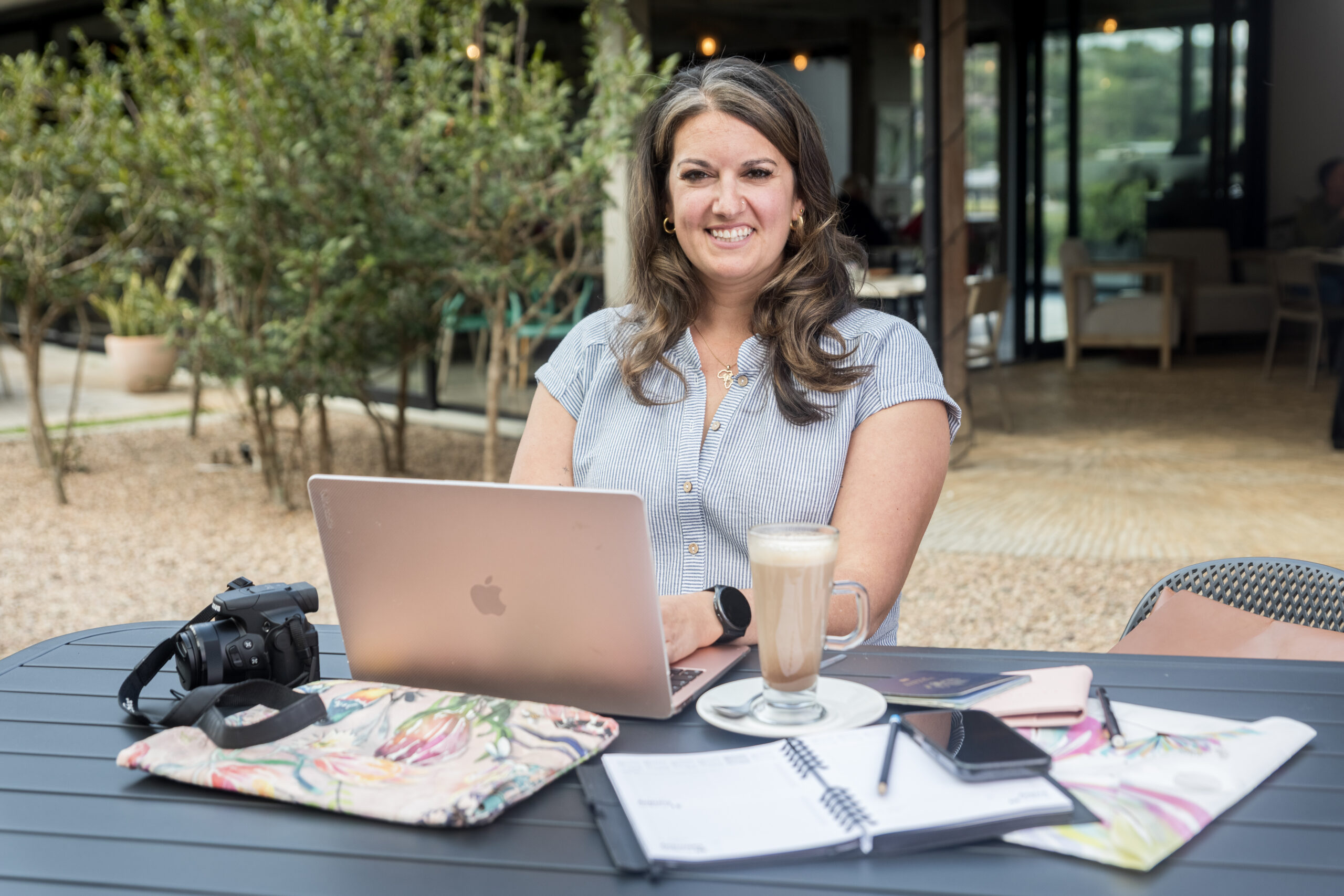 Baylee Shapiro a Luxury Travel Advisor of BTA working remotely at a stylish outdoor café with laptop, camera, and planner.